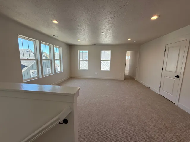 Spacious upper-level room featuring light-colored carpeting, recessed lighting, multiple windows, a white paneled door, and white painted trim