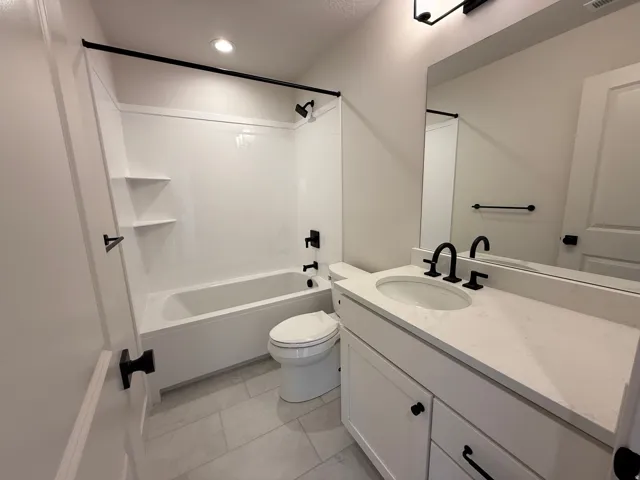 Full bathroom featuring a white tub and shower surround with built-in shelving, matte black fixtures, a single vanity with a solid surface countertop, and neutral-toned floor tiling