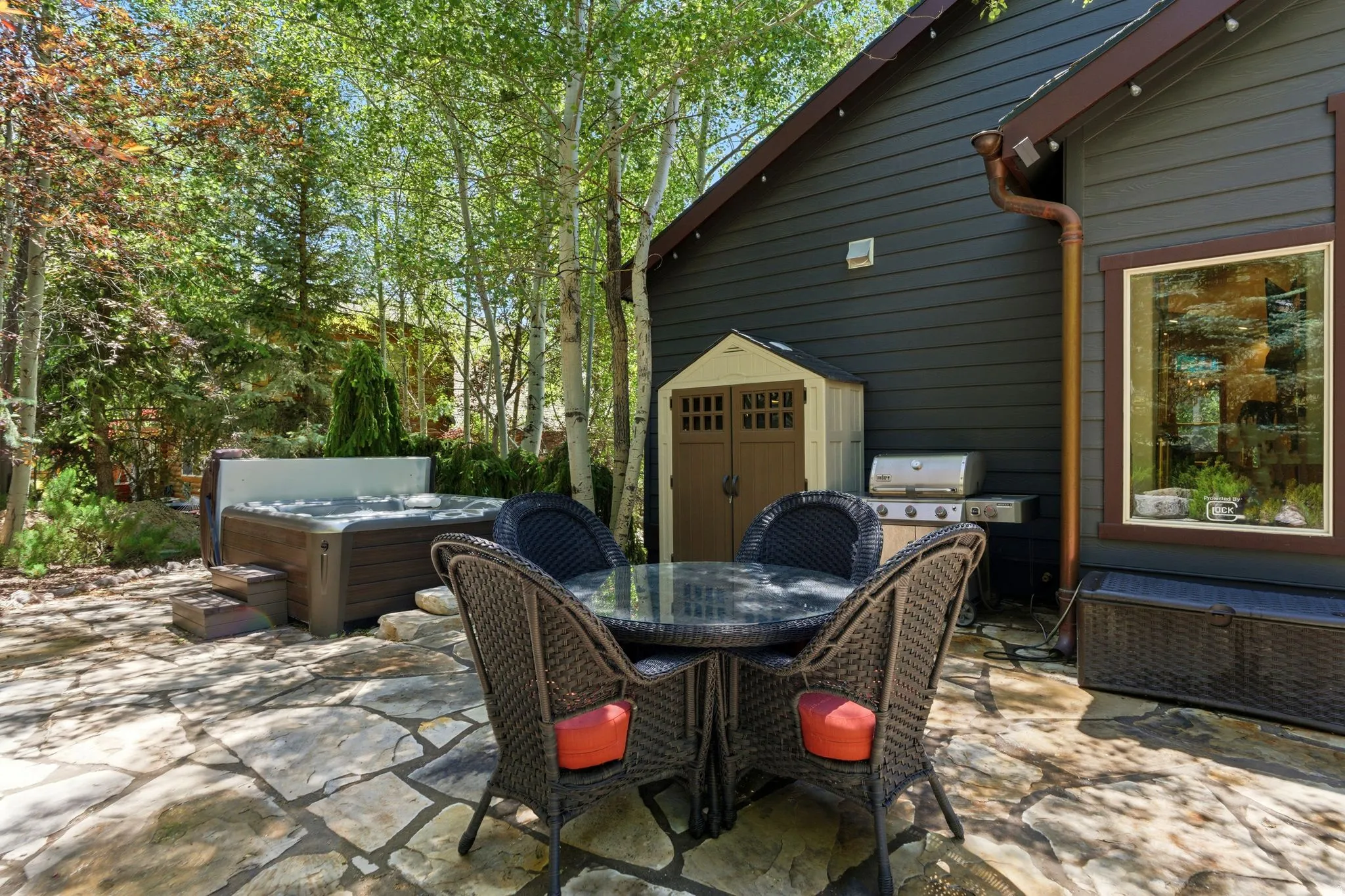 Stone patio area featuring a built-in hot tub with steps, a dark-paneled exterior, and mature surrounding trees