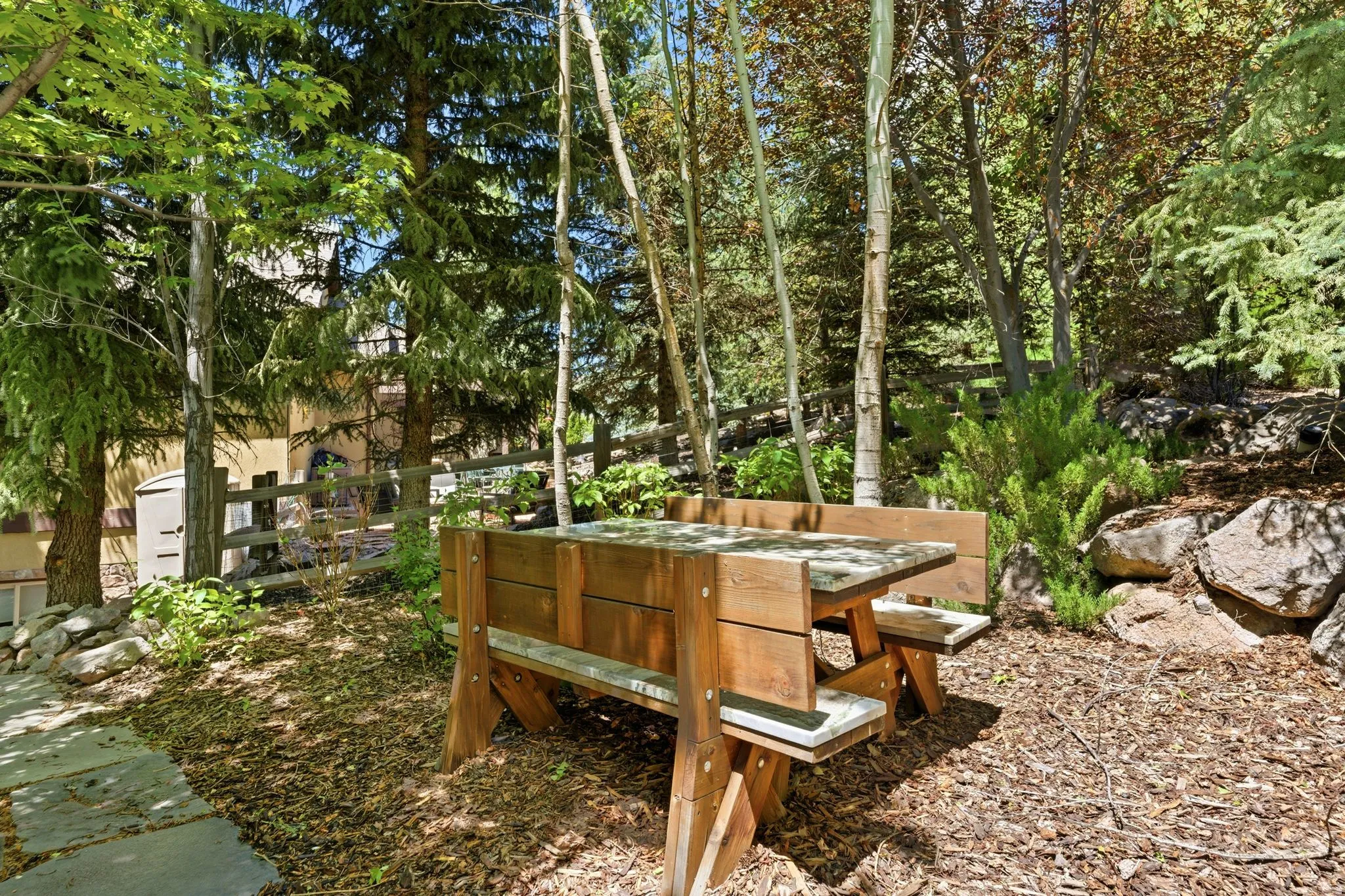 Outdoor dining area featuring a wooden picnic table with benches, surrounded by diverse mature trees and natural rock landscaping