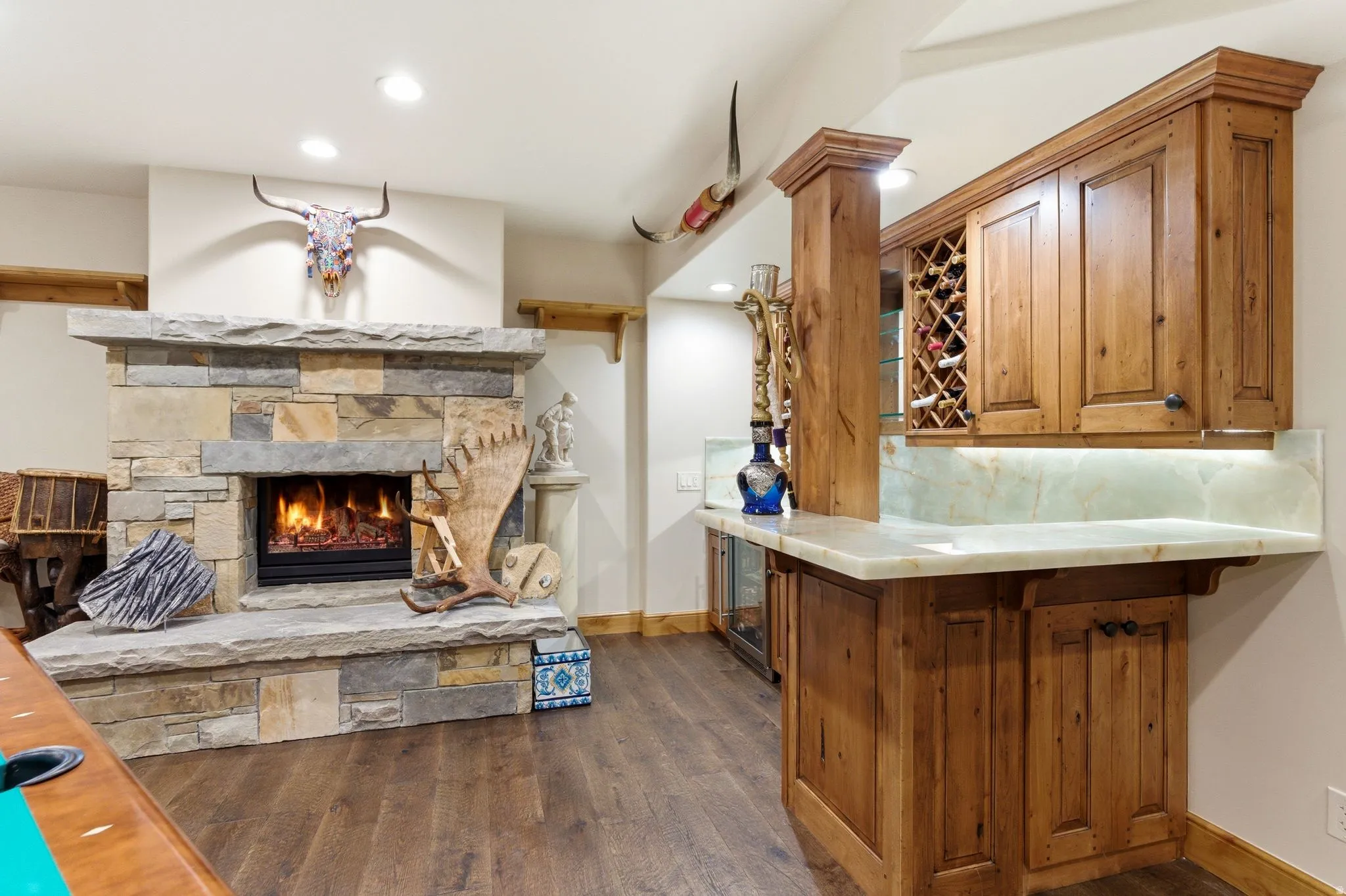 Stone-faced fireplace with a substantial hearth, wood-finish flooring, and a built-in wet bar featuring wood cabinetry, a wine rack, and a light-toned countertop