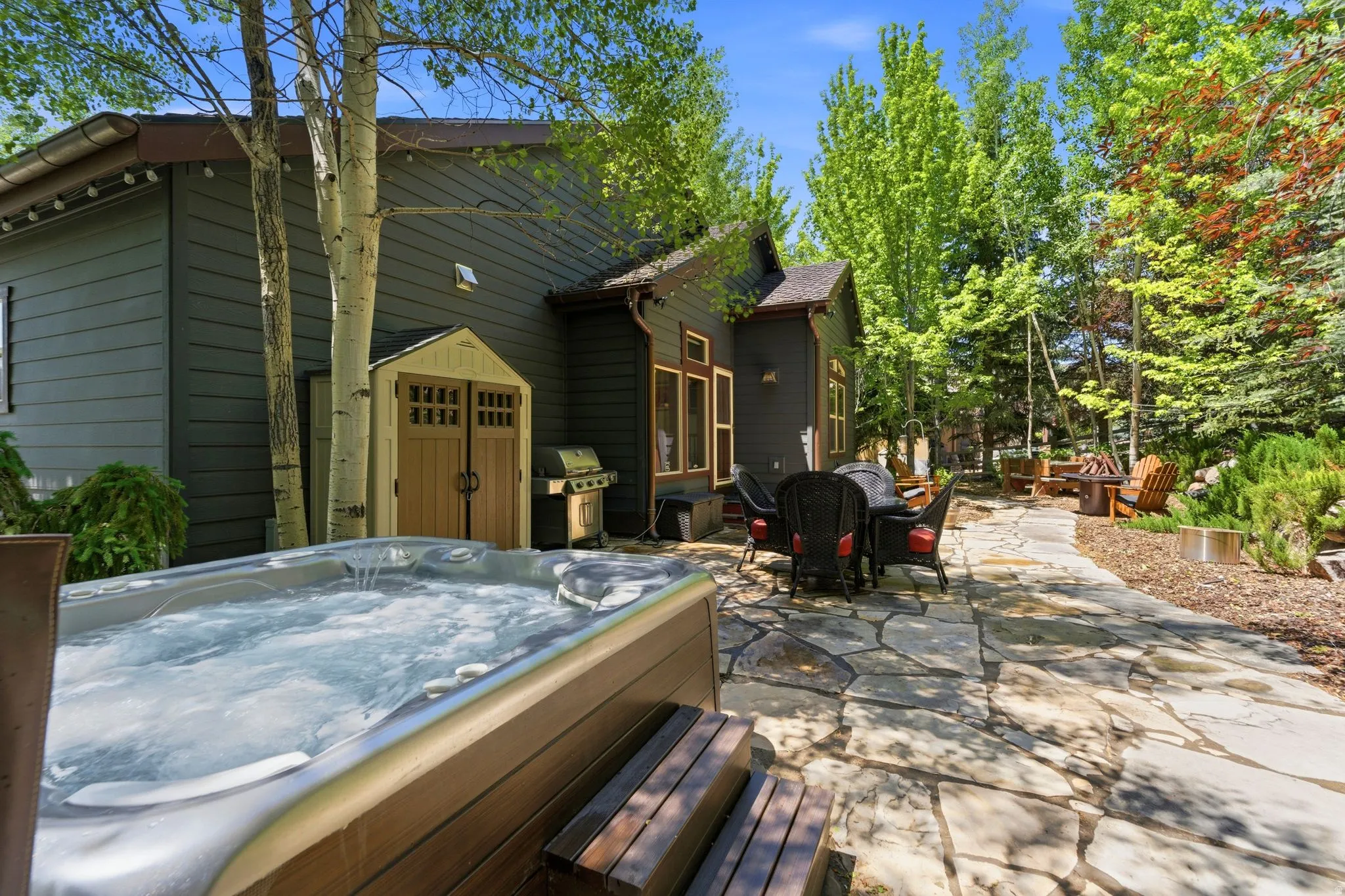 Outdoor spa with wood-finish surround, extensive flagstone patio, outdoor dining area, mature trees, and a storage shed