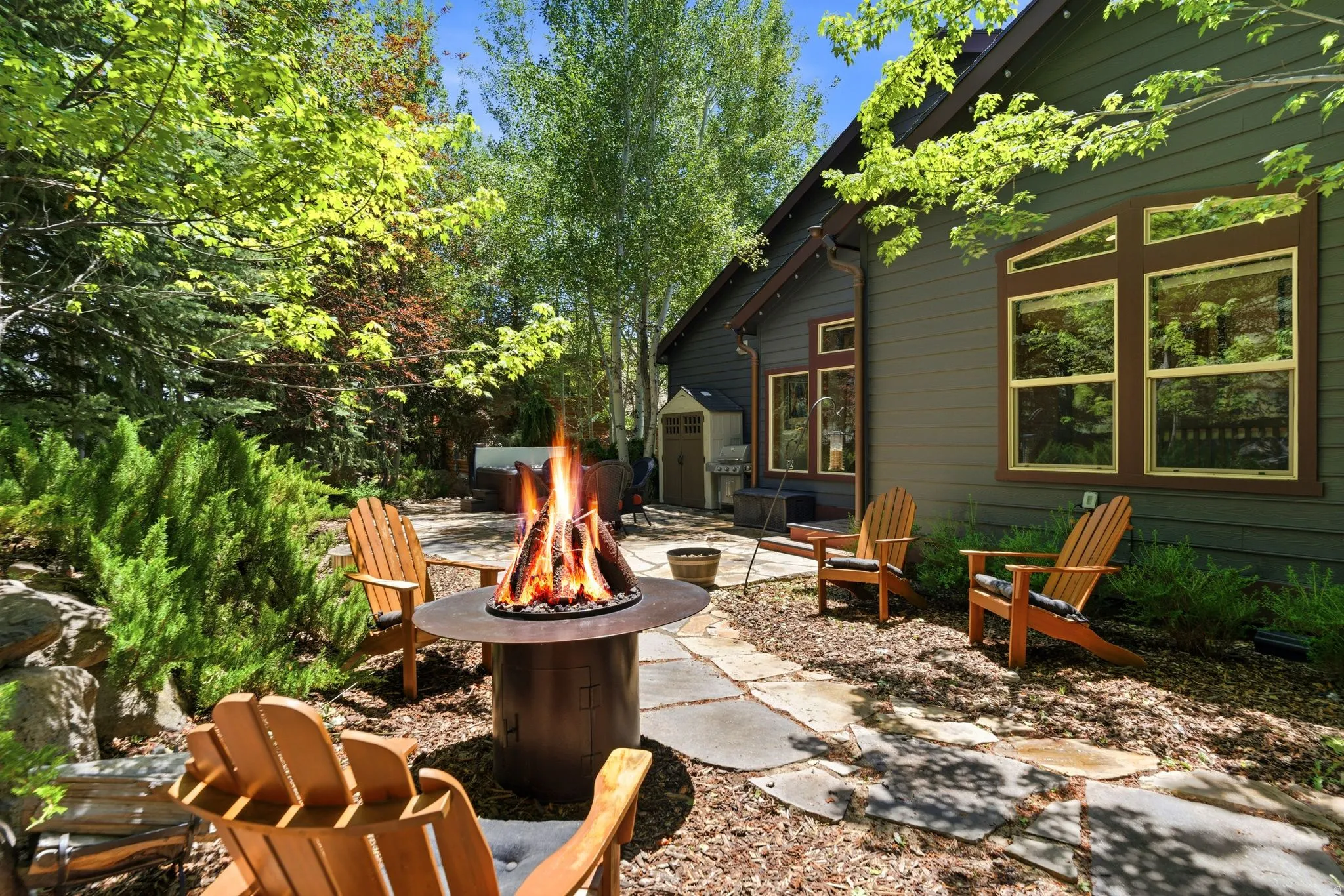 Stone patio area featuring a built-in fire pit, surrounded by lush landscaping and mature trees