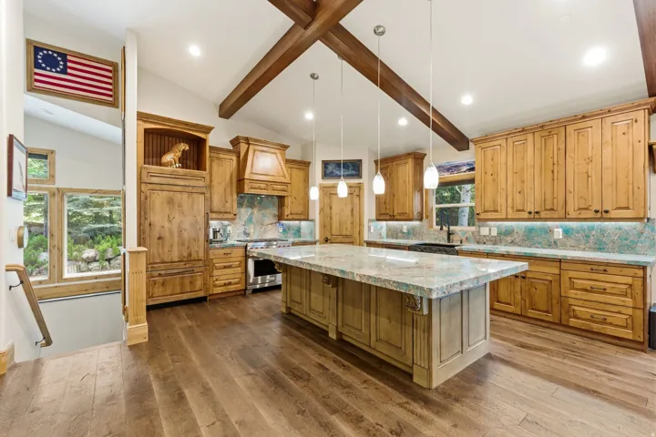 Spacious kitchen featuring vaulted ceilings with exposed wood beams, wood-finish flooring, custom wood cabinetry, a large center island with a light-toned countertop, and a tiled backsplash