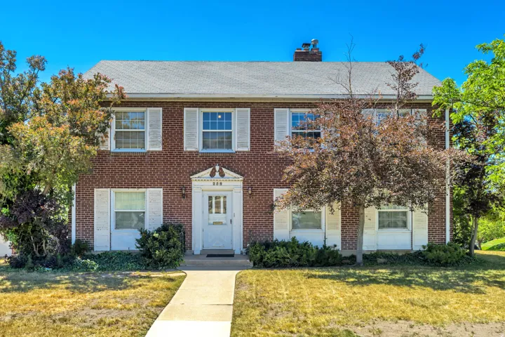 Brick exterior property featuring a symmetrical facade, white decorative window shutters, a white paneled front door, a concrete pathway, and mature landscaping