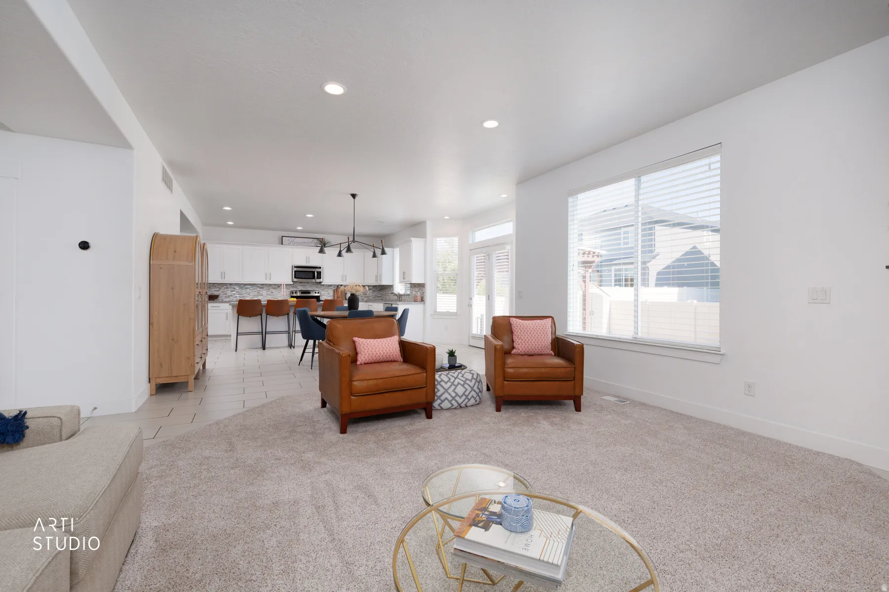 Spacious open-concept living area featuring light-toned carpeting, ample natural light from large windows, recessed lighting, and a kitchen with white cabinetry and a dark backsplash