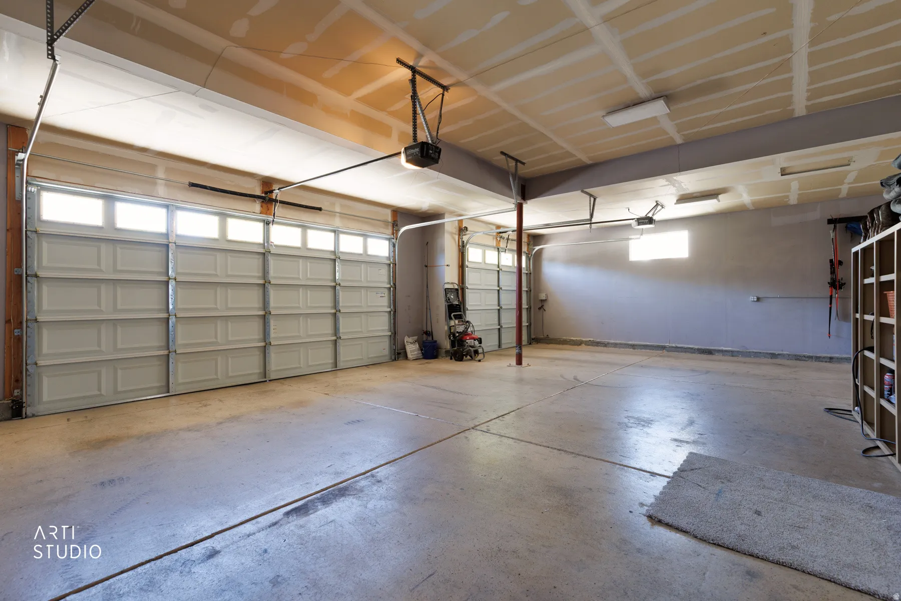 Spacious multi-car garage featuring two overhead doors with transom windows, polished concrete flooring, exposed ceiling beams, and a built-in shelving unit