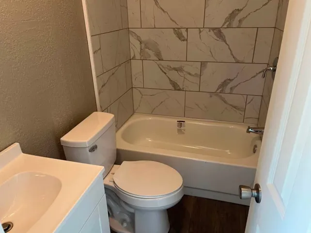 Full bathroom featuring a white bathtub with tile surround, a white toilet, a vanity with an integrated sink, and wood-finish flooring