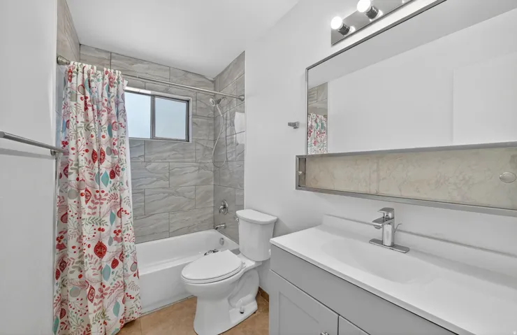 Contemporary bathroom featuring a tub/shower combination with grey tile surround, a built-in window, a vanity with a white countertop and integrated sink, and an overhead mirror with integrated lighting