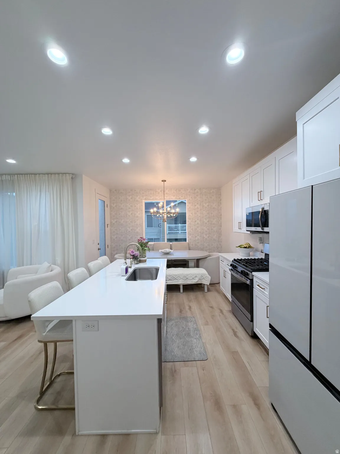 Contemporary open-concept kitchen featuring light wood-finish flooring, a spacious white island with an integrated sink, white cabinetry, and stainless steel appliances including a gas range and French door refrigerator