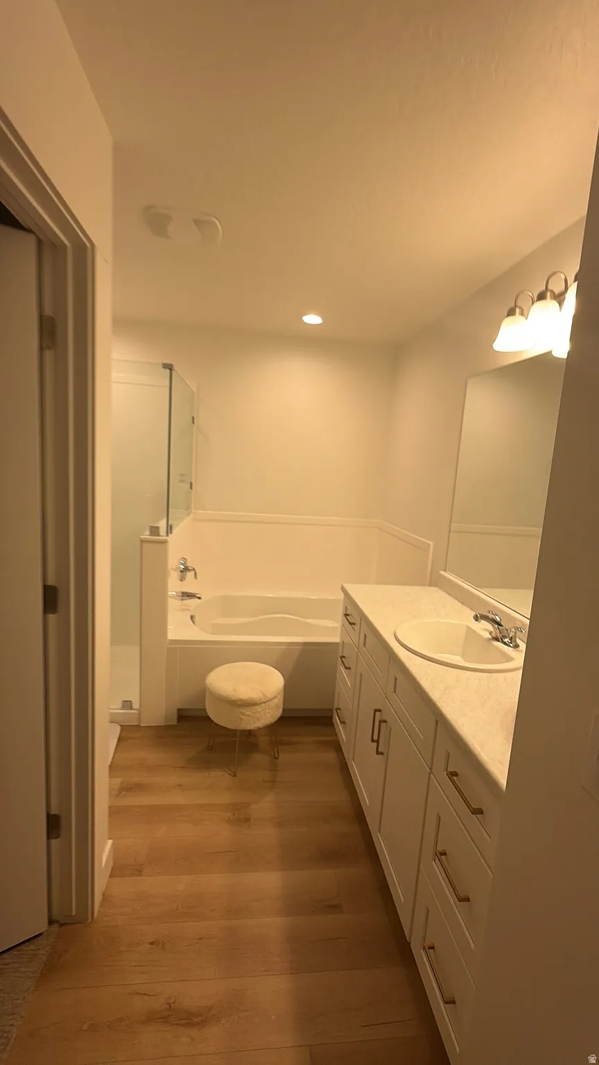Bathroom featuring a double vanity with white cabinetry and integrated sinks, a built-in bathtub, a walk-in shower with a glass enclosure, and wood-finish flooring