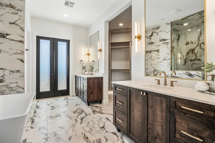 Spa-inspired bathroom featuring large format marble-look tile flooring and accent wall, dual dark wood vanities with white countertops, and a built-in walk-in closet with shelving