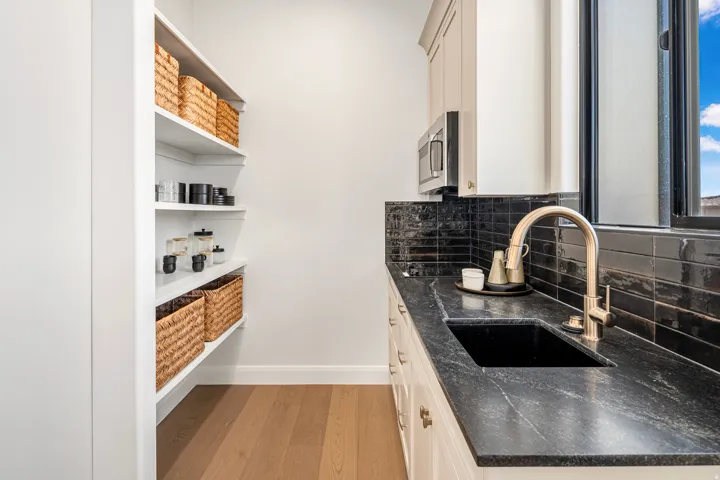 Modern pantry featuring extensive open shelving, wood-finish flooring, a deep black sink, a stainless steel microwave, and a dark stone countertop with a sleek tile backsplash