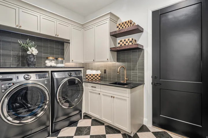Dedicated utility room featuring abundant cabinetry, a dark tile backsplash, a built-in sink with a bronze-finish faucet, and a contrasting black and white checkered floor
