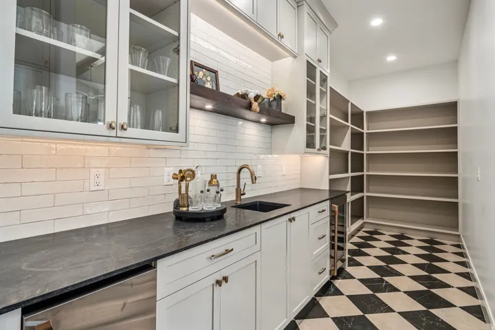 Walk-in pantry featuring extensive built-in shelving, black and white checkered flooring, a wet bar with a dark stone countertop, white subway tile backsplash, and upper cabinetry with glass fronts