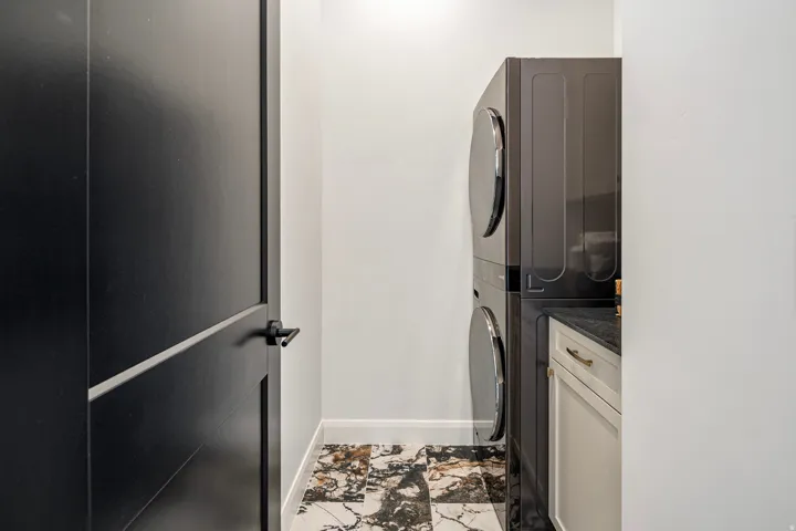 Laundry area featuring a stacked washer and dryer, black countertop, light cabinetry with gold-tone hardware, and patterned floor tiling
