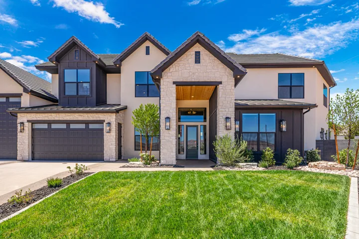 Striking two-story residence featuring a stone and stucco exterior, a prominent covered entry with wood-finish ceiling, multiple large windows with dark framing, a two-car garage with overhead lighting, and a well-maintained front lawn