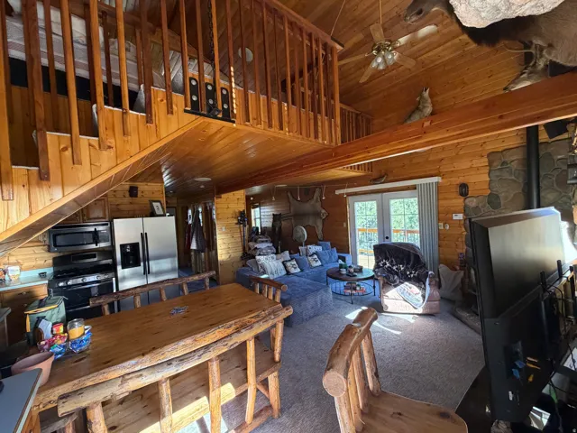 Open-concept living area with wood-finish walls and ceilings, a wooden staircase leading to an upper loft, and a stone-finish fireplace