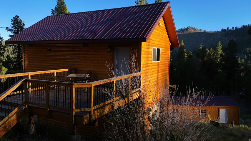 Log cabin exterior featuring a reddish-brown metal roof, an elevated wooden deck with black metal balusters, and a natural wooded backdrop