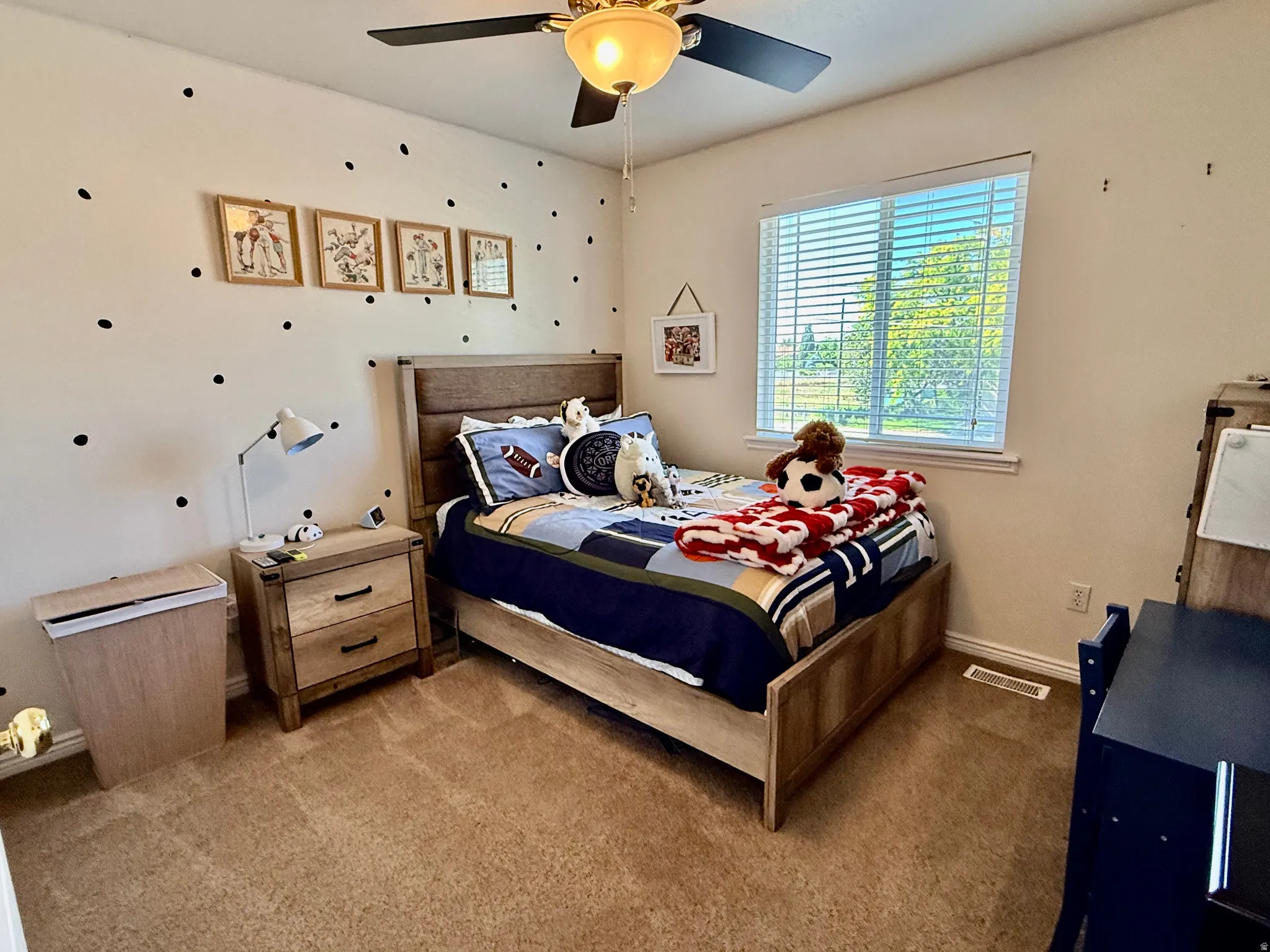 Carpeted bedroom featuring a ceiling fan with light fixture, a large window with blinds, and a built-in air vent