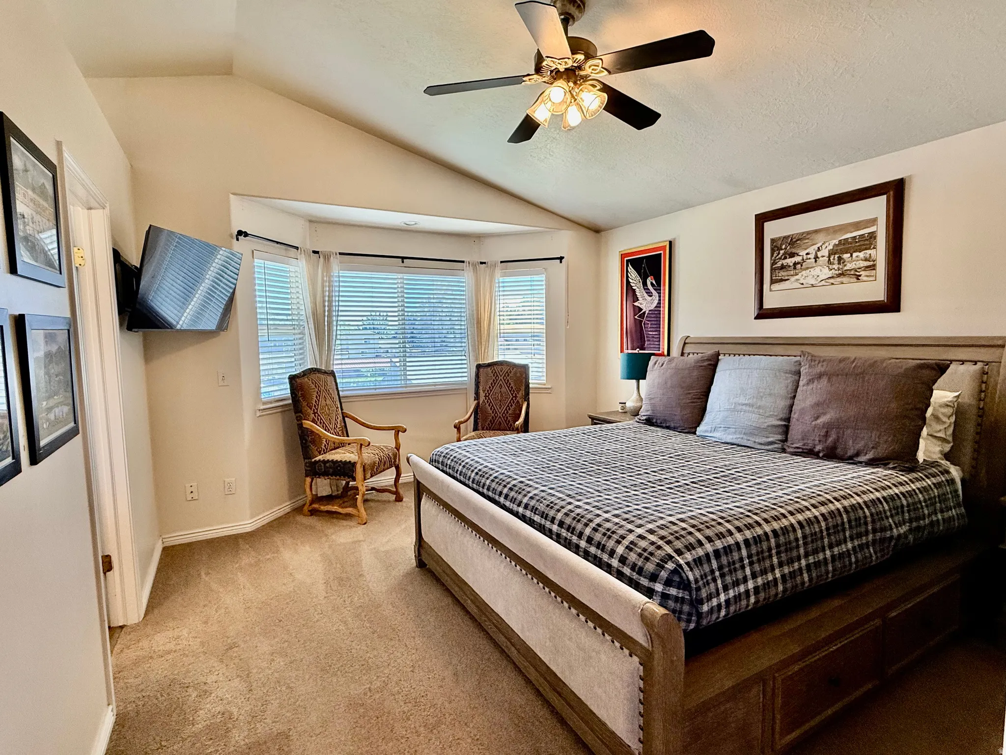 Vaulted ceiling bedroom featuring a bay window with blinds, a ceiling fan with integrated lighting, a wall-mounted television, and carpet flooring