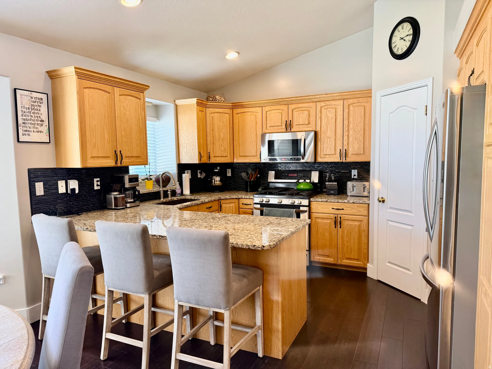 Kitchen featuring wood-finish cabinetry, granite countertops, a dark tile backsplash, stainless steel appliances, and dark wood-finish flooring