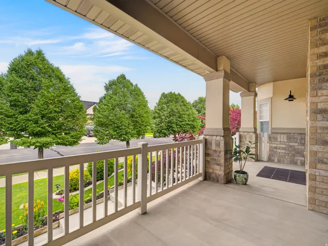 Covered porch featuring concrete flooring, stone-clad columns, and a railing overlooking a landscaped front yard with mature trees