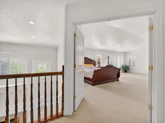 Expansive bedroom featuring neutral carpeting, a tray ceiling, recessed lighting, multiple windows with plantation shutters, and a classic wood baluster railing visible from the entryway