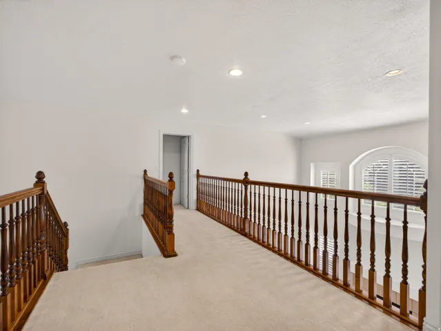 Upper-level landing featuring a wood-finish banister, neutral carpet flooring, recessed lighting, and an arched window with plantation shutters