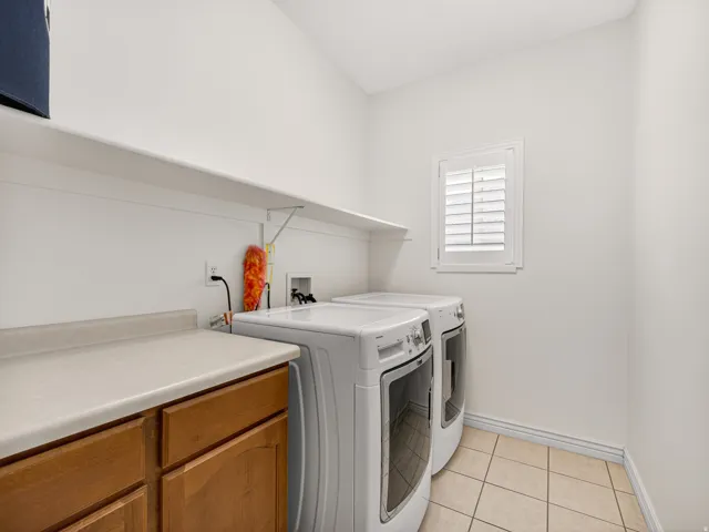 Dedicated laundry area featuring a window with white louvered shutters, tiled flooring, wooden cabinetry with a light-toned countertop, and a white wall-mounted shelf