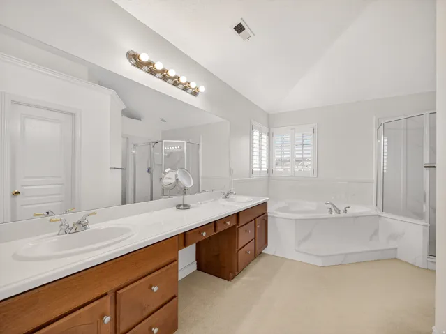 Spacious bathroom featuring a dual vanity with wood cabinetry, a built-in makeup vanity, an integrated soaking tub with stone-look surround, a separate glass-enclosed shower, and plantation-shuttered windows