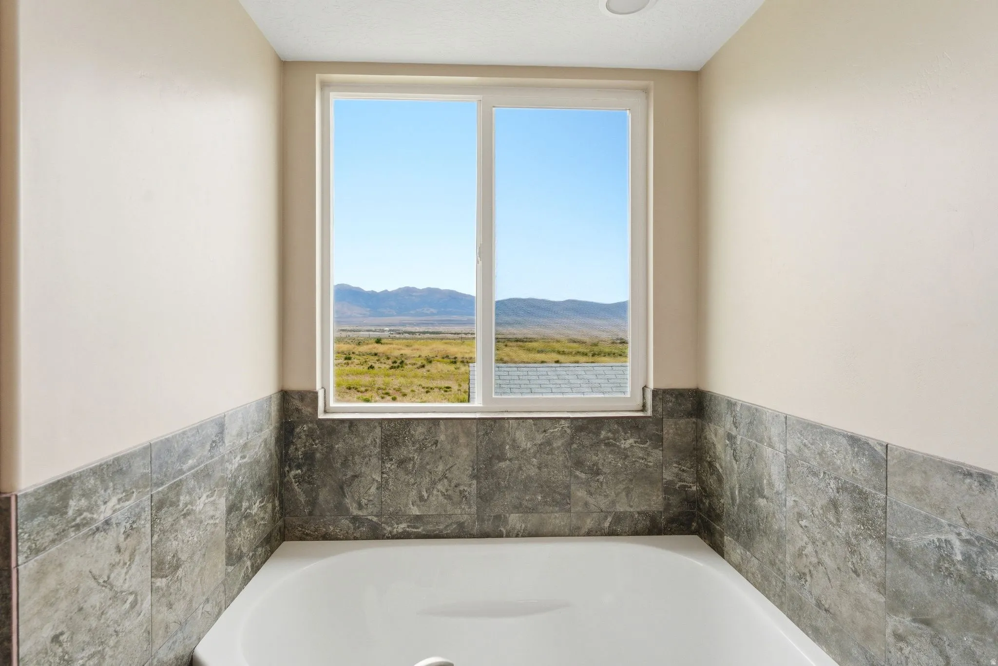 Built-in soaking tub with gray marble-look tile surround and a large window presenting a distant mountain and field landscape