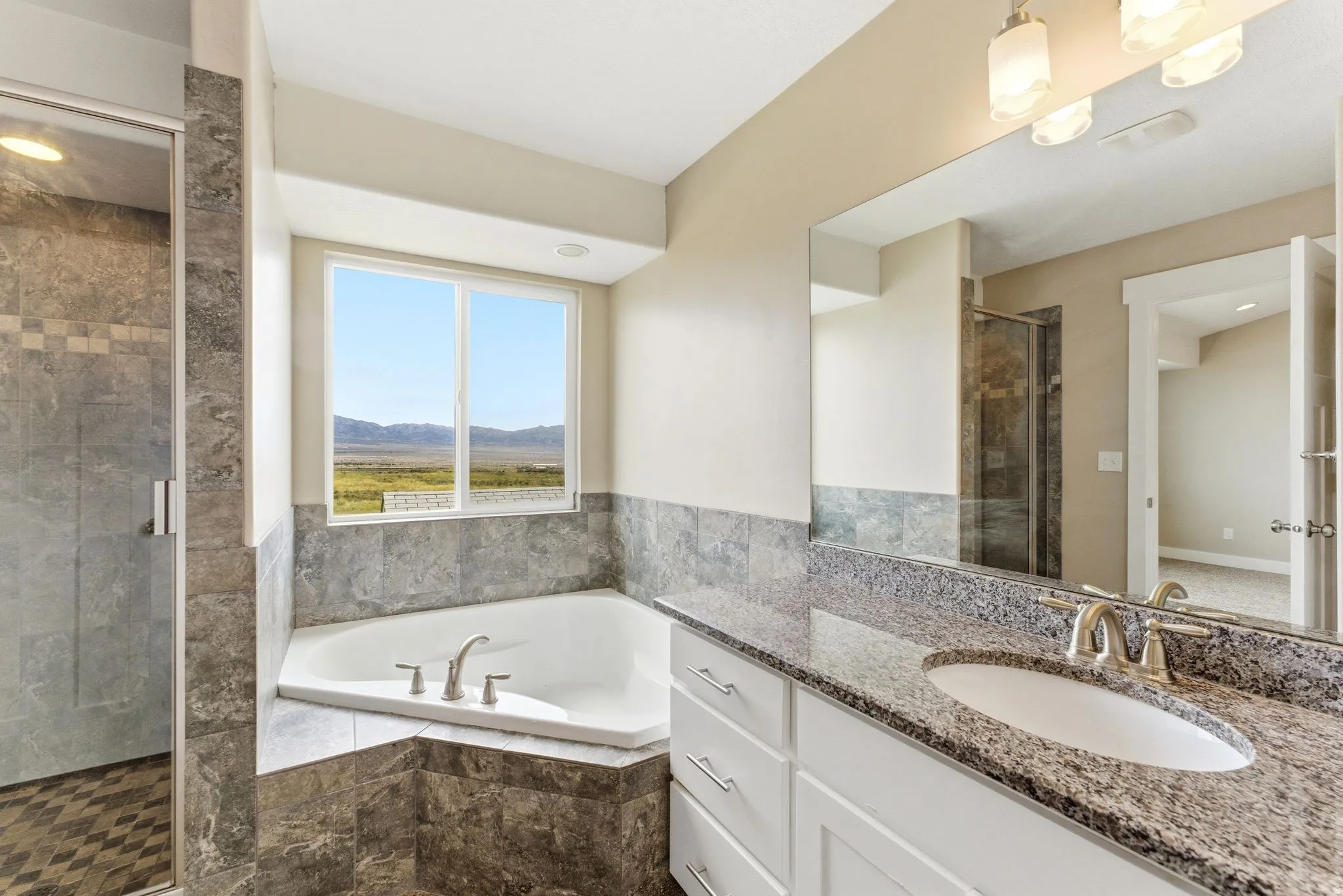 Corner soaking tub with a tiled surround, a separate glass-enclosed shower, a double vanity with a stone-finish countertop, and a large window