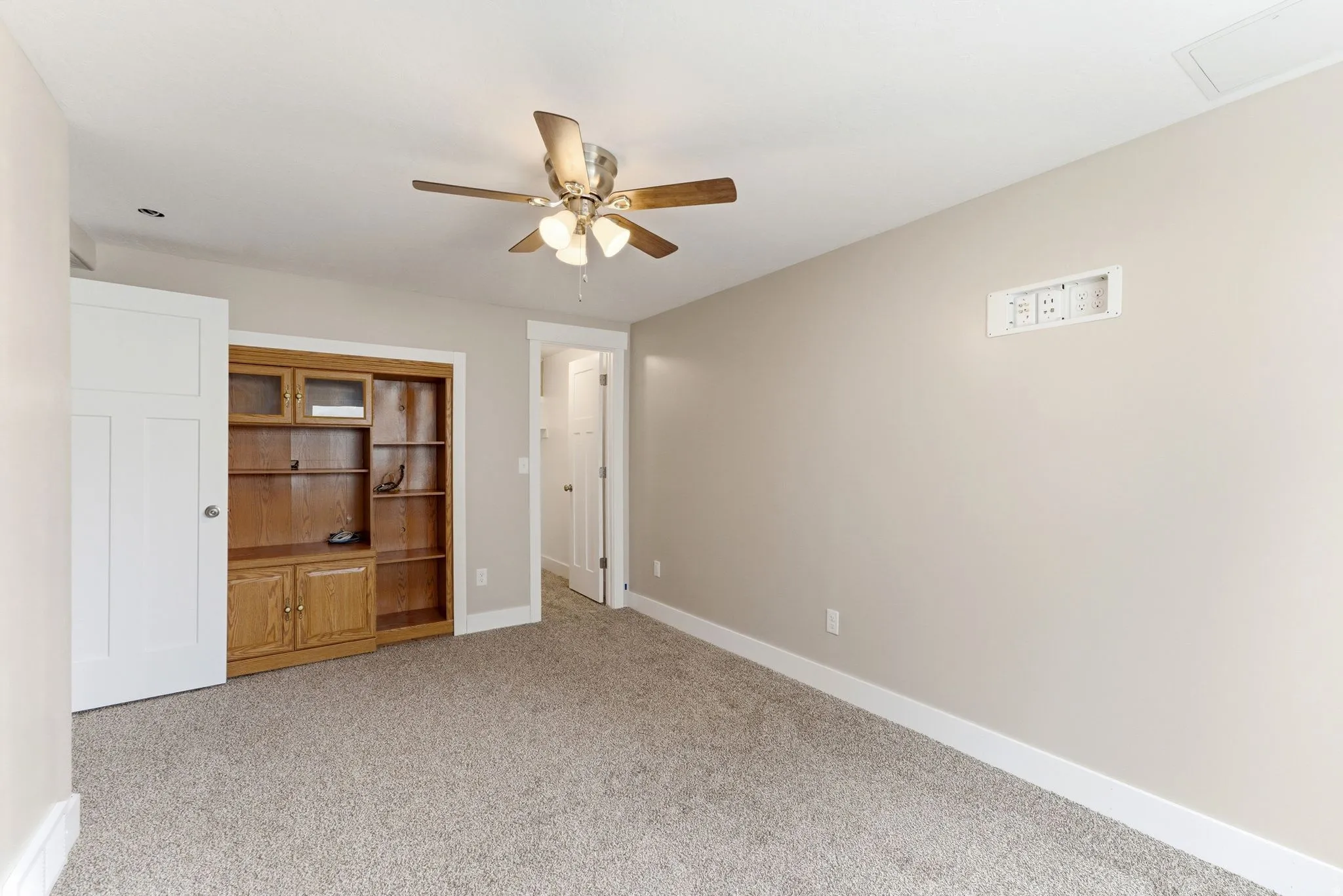 Neutral-toned carpeted room featuring a built-in wood-finish shelving unit, a ceiling fan with integrated lighting, and white baseboards