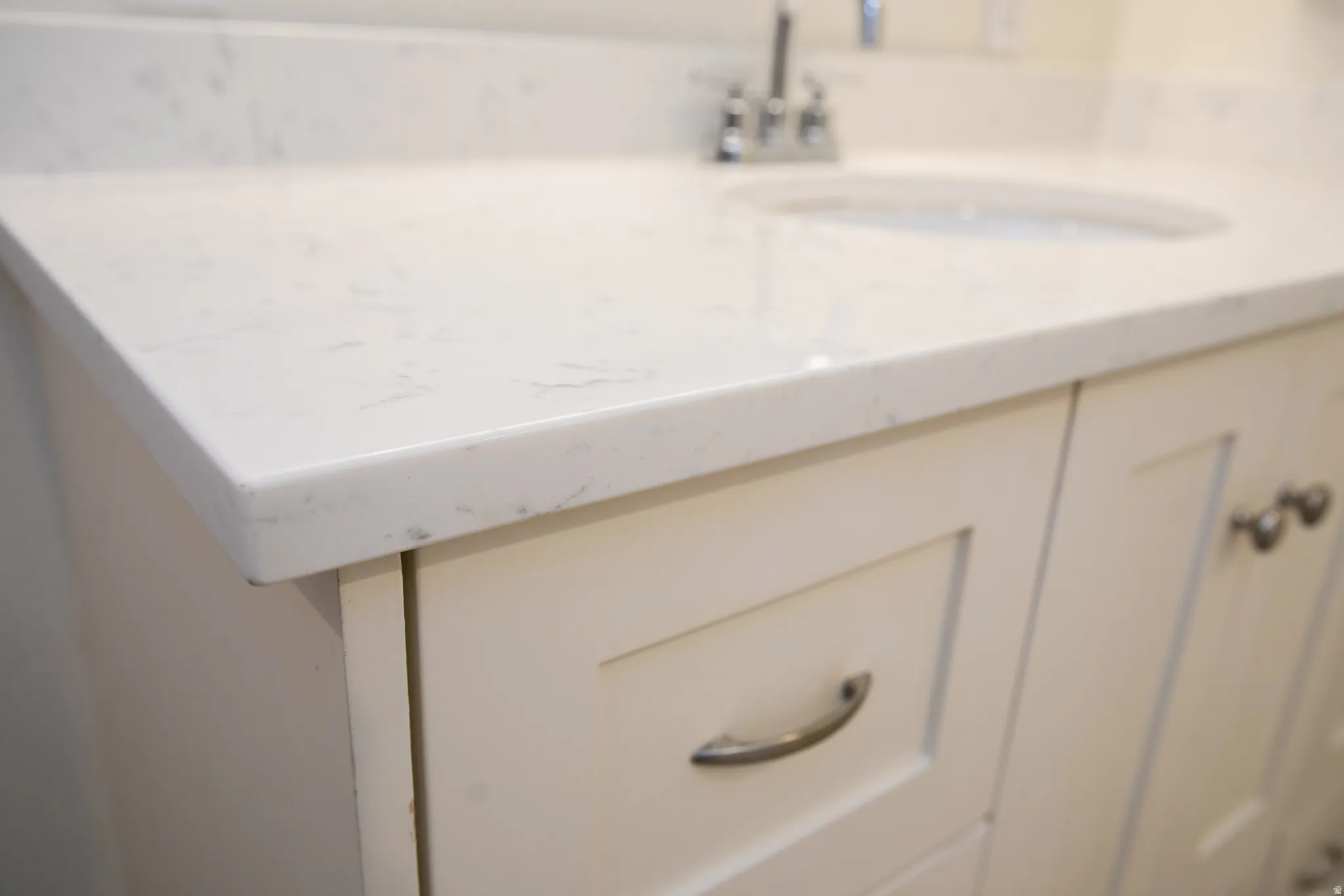 White countertop with subtle veining, undermount sink, chrome faucet, and off-white shaker-style cabinetry with brushed nickel hardware