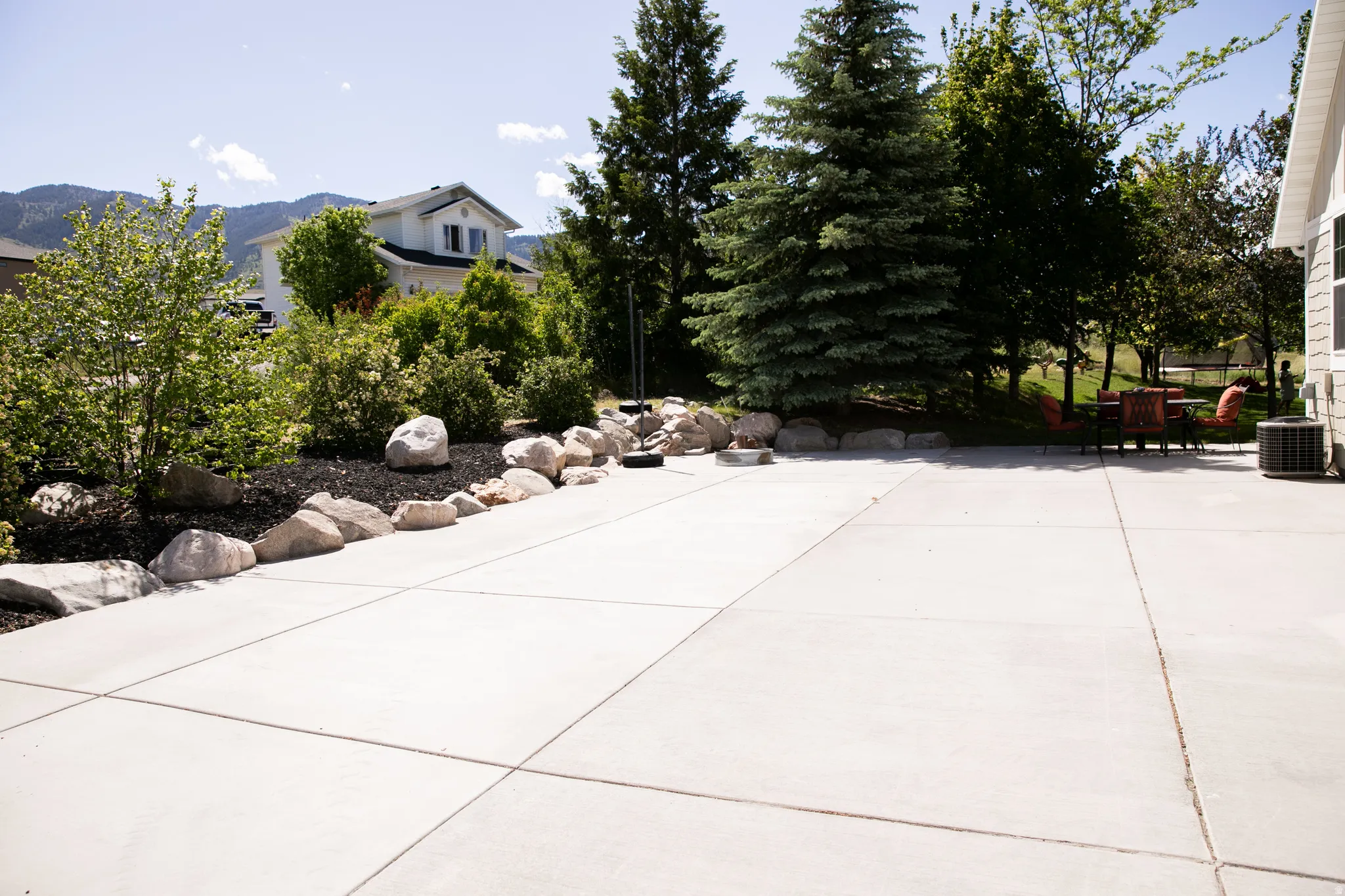 Expansive concrete patio with integrated landscaping featuring large natural boulders, dark mulch, and mature trees