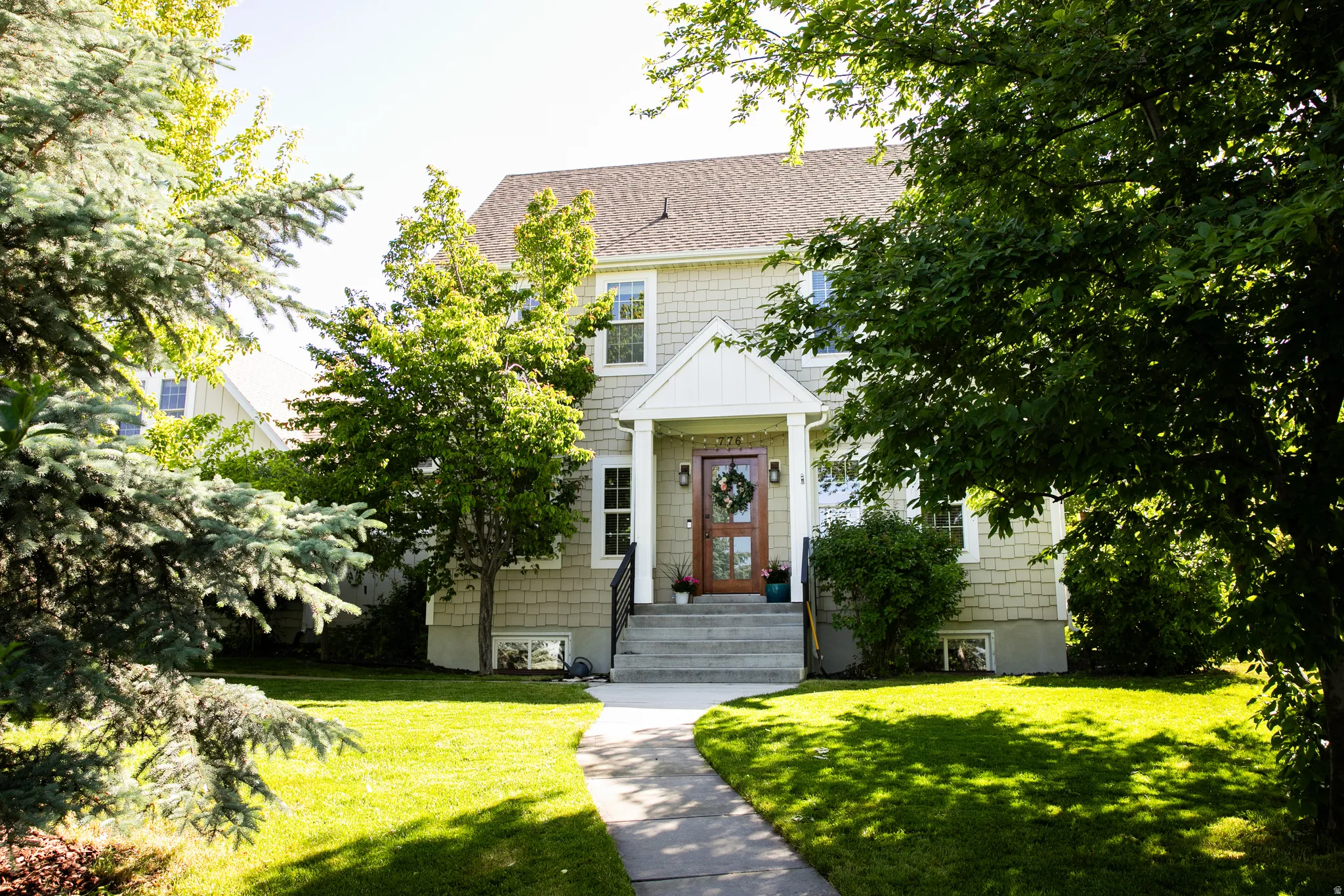 Shingle-sided residence with gabled entry porch, wood-finish front door, concrete walkway, and mature trees