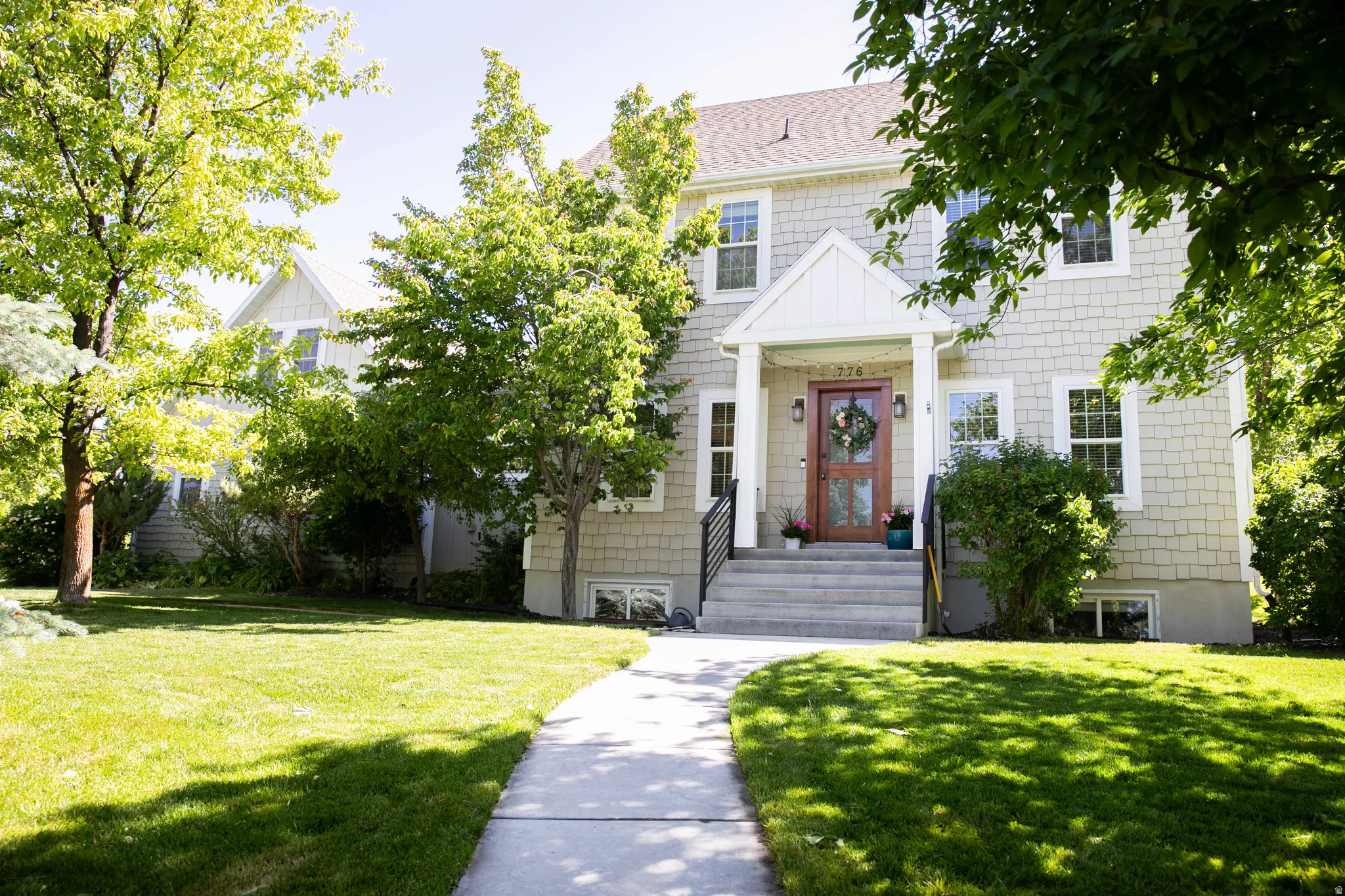 Shingle-sided residence featuring a prominent gabled entry, concrete steps with black iron railings, a wood-finish front door, and a concrete pathway through a grassy lawn with mature trees