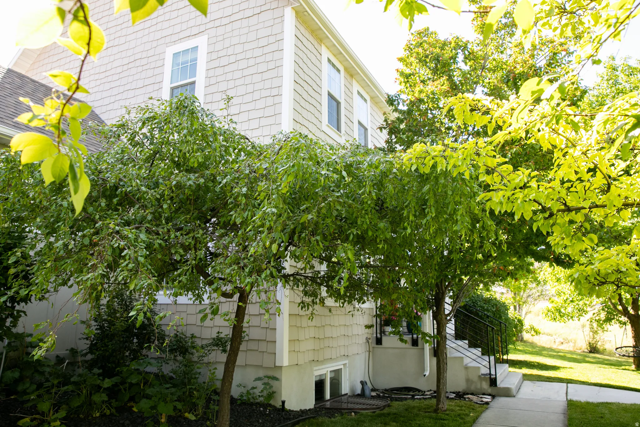Exterior featuring shingle siding, multiple mature trees, a concrete pathway, and steps with a metal handrail