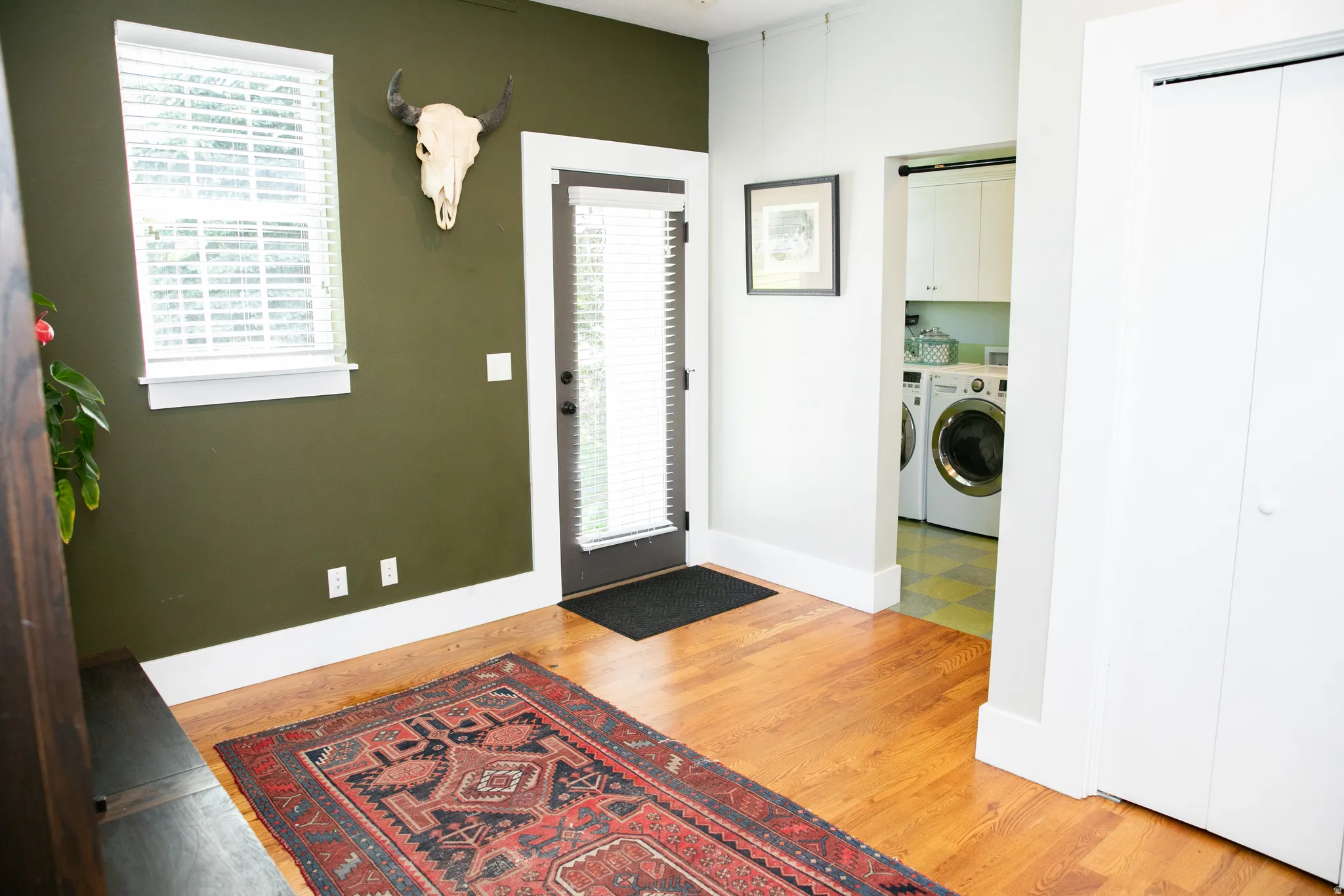 Rich hardwood flooring throughout, featuring a window with blinds, a dark-framed exterior door, and a dedicated laundry area with built-in cabinetry and tile flooring