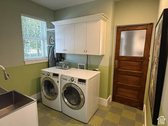 Main-floor laundry.  Checkered flooring, a window with horizontal blinds, overhead white cabinetry, and a utility sink with a pull-down faucet