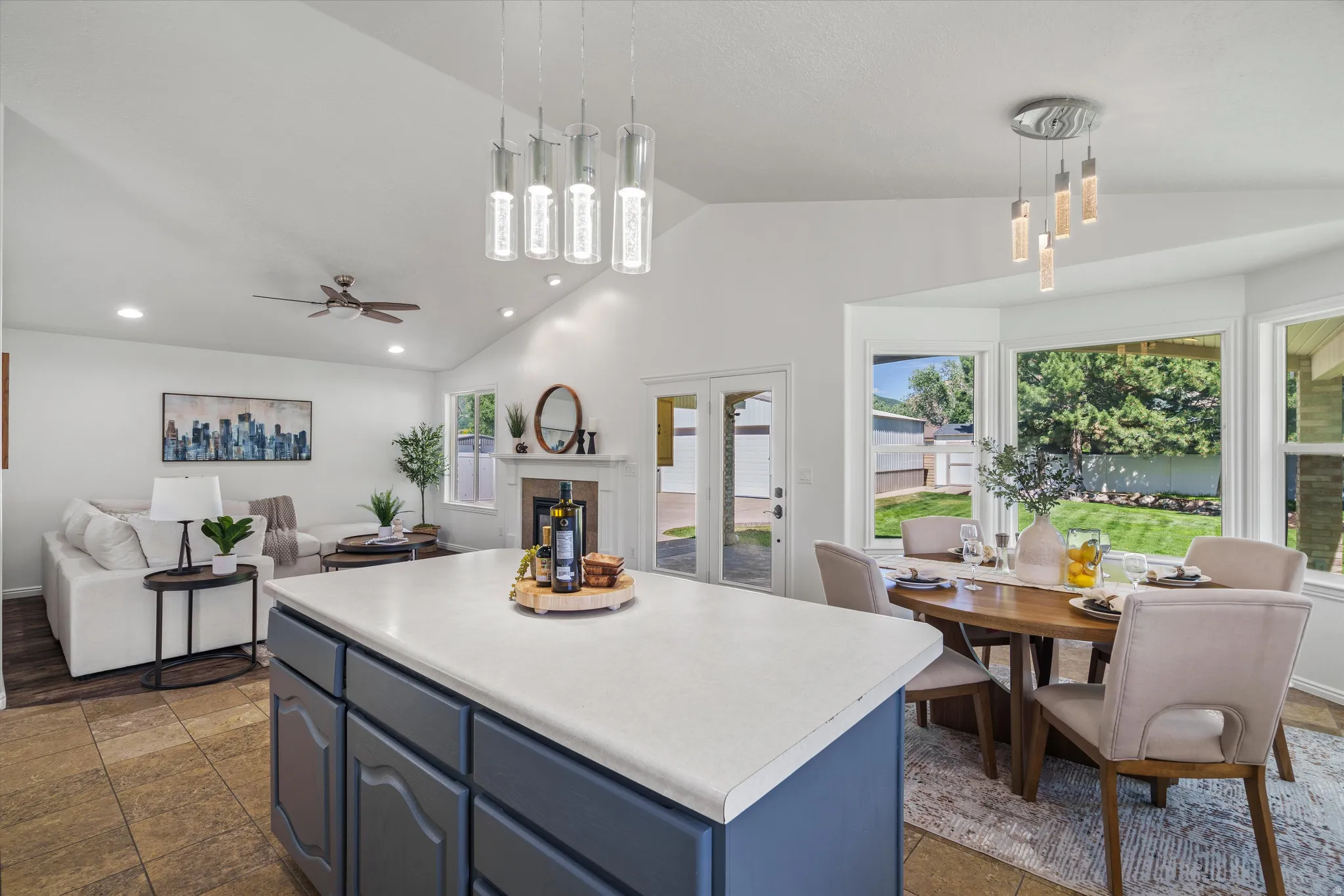 Open-concept living area featuring a kitchen island with a light-colored countertop, recessed lighting, vaulted ceilings, a ceiling fan, and a dining area with a bay window overlooking a backyard