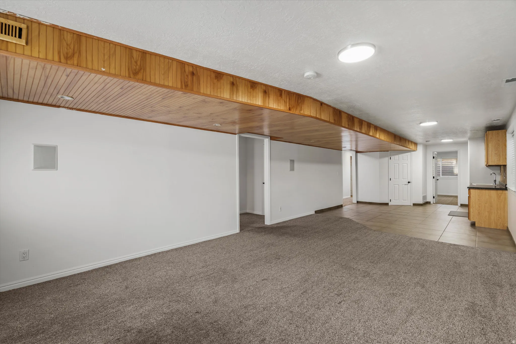 Spacious lower-level area featuring a carpeted main zone, tiled flooring, wood-paneled ceiling accents, and a built-in wet bar with wood-finish cabinetry