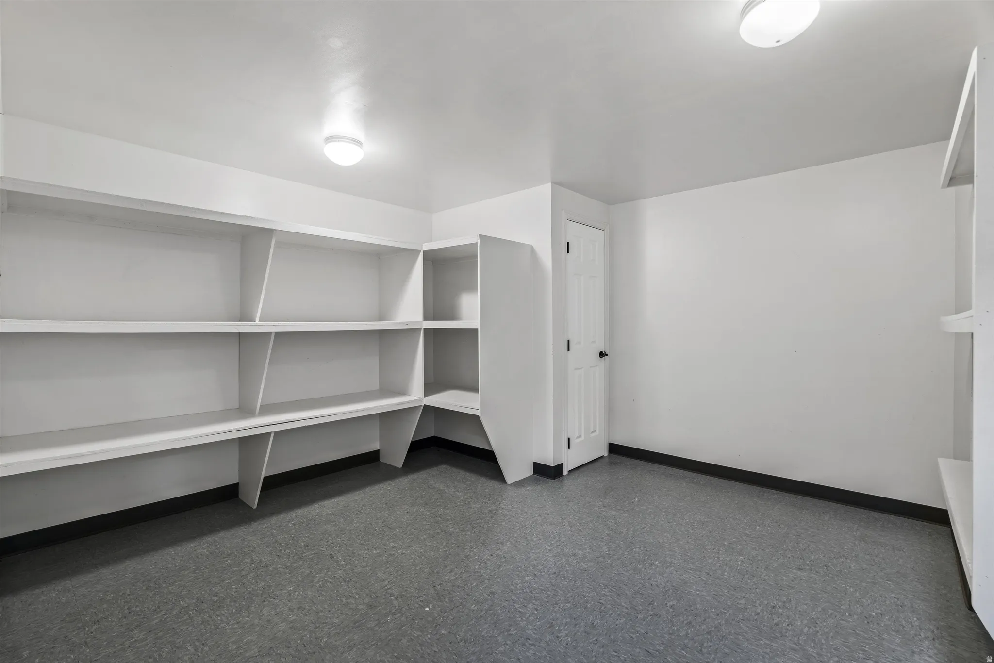 Dedicated storage room featuring extensive built-in shelving, durable gray flooring, crisp white walls, and overhead lighting. Located at the bottom of the staircase across from the kitchenette.