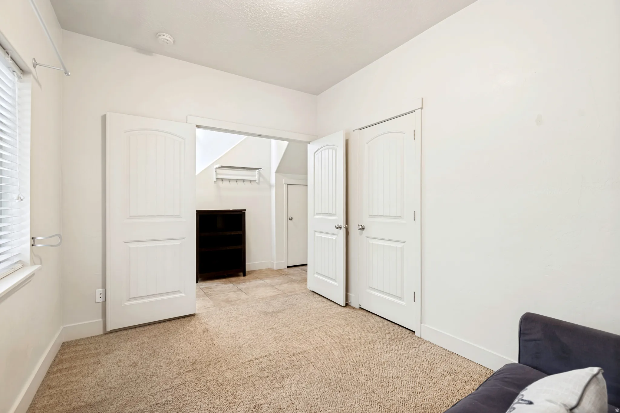 Carpeted room with white walls, featuring a window with blinds, two white paneled doors, and an open doorway leading to a tiled area with a dark built-in shelving unit