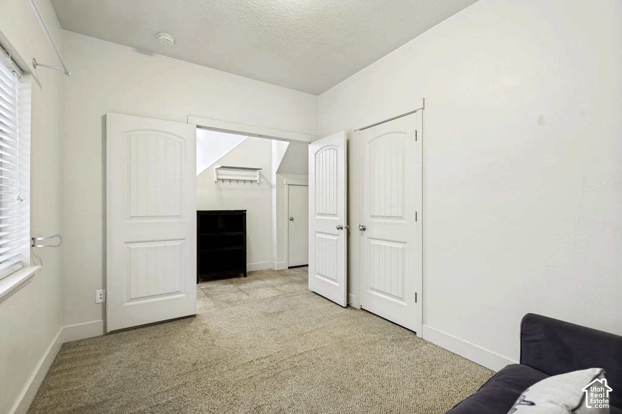 Carpeted room with white walls, featuring a window with blinds, two white paneled doors, and an open doorway leading to a tiled area with a dark built-in shelving unit