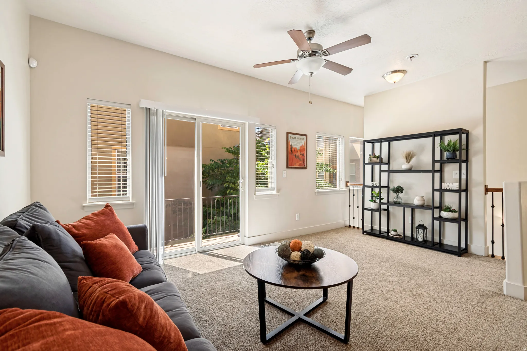 Living area featuring neutral wall tones, textured carpeting, a sliding glass door leading to a balcony, multiple windows with blinds, and a permanent ceiling fan with integrated lighting