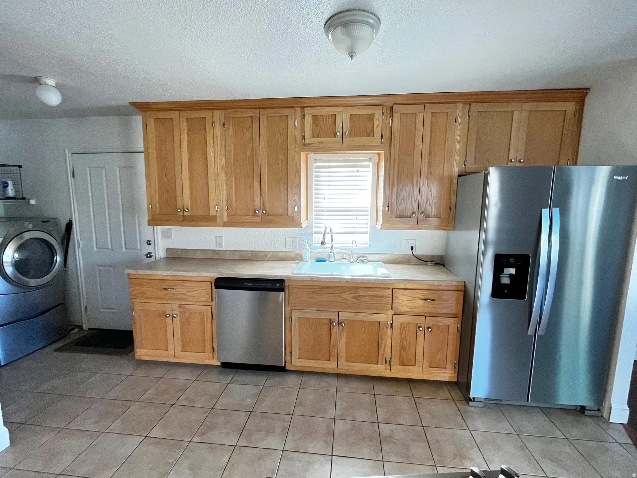 Kitchen featuring wood cabinetry, light-toned countertops, a stainless steel dishwasher, a stainless steel side-by-side refrigerator, and tile flooring