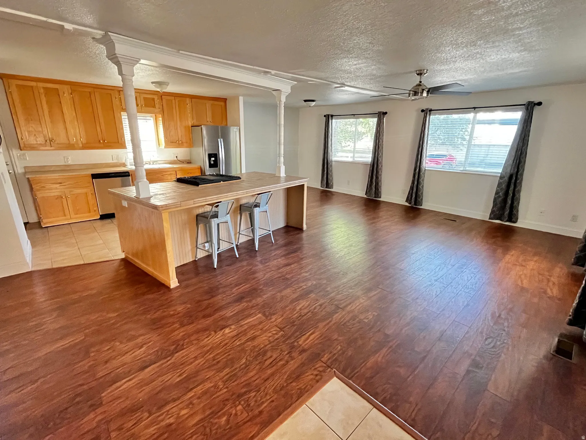 Open-concept living space featuring wood-finish flooring, a kitchen island with seating, natural wood cabinetry, stainless steel appliances, and a ceiling fan