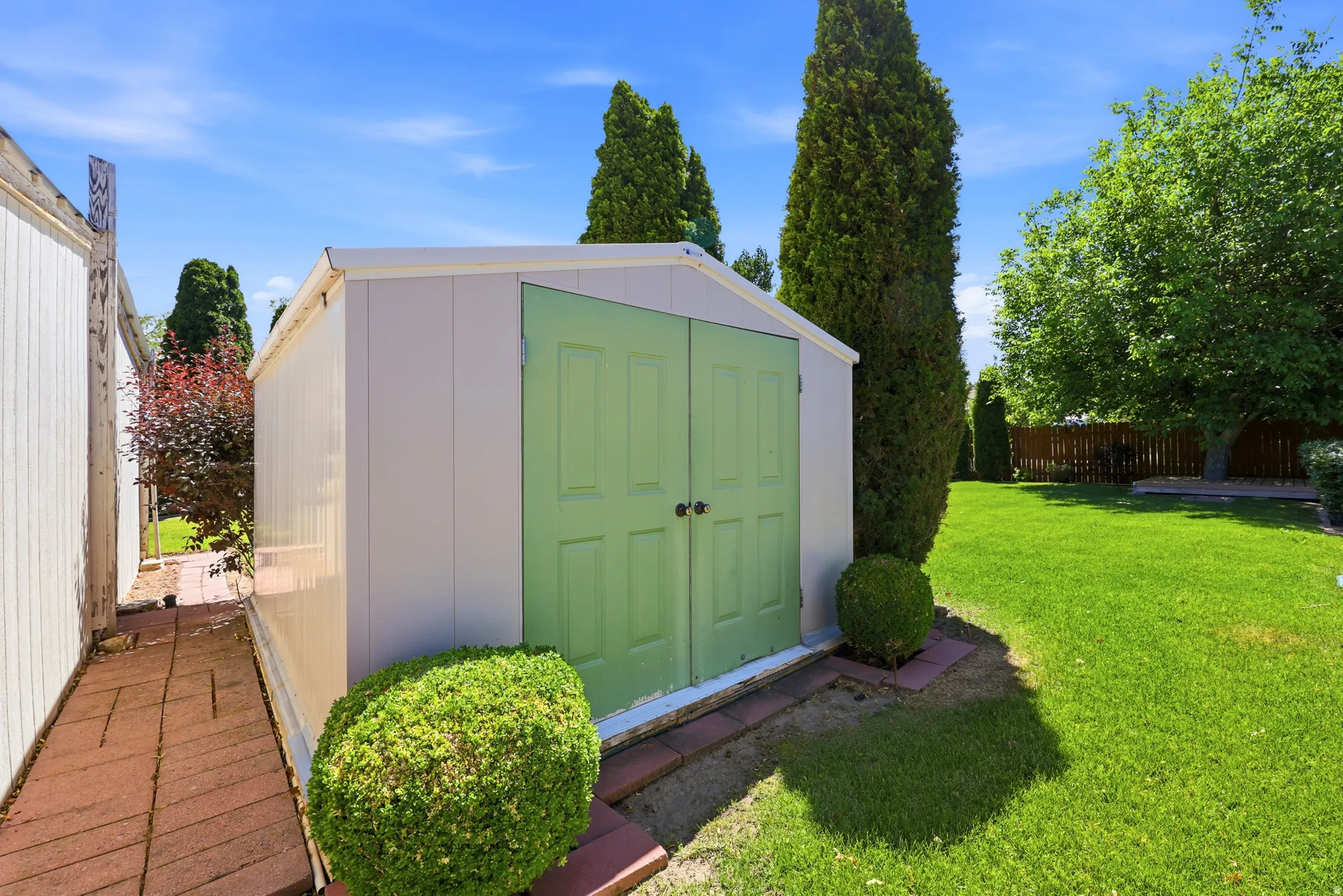 Detached storage shed with light green double doors, bordered by a manicured shrub and brick pavers